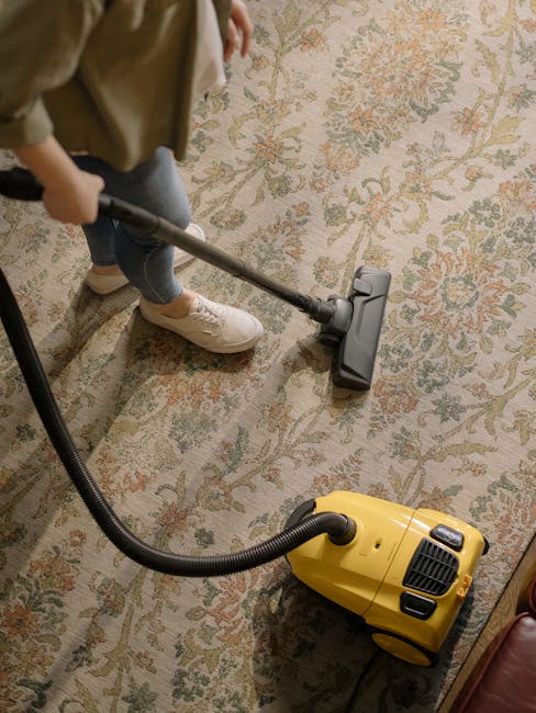 A professional cleaner wearing a black glove is using a handheld steam or extraction machine to deep clean a dark gray carpeted floor in a commercial or residential setting. The machine has a white ta